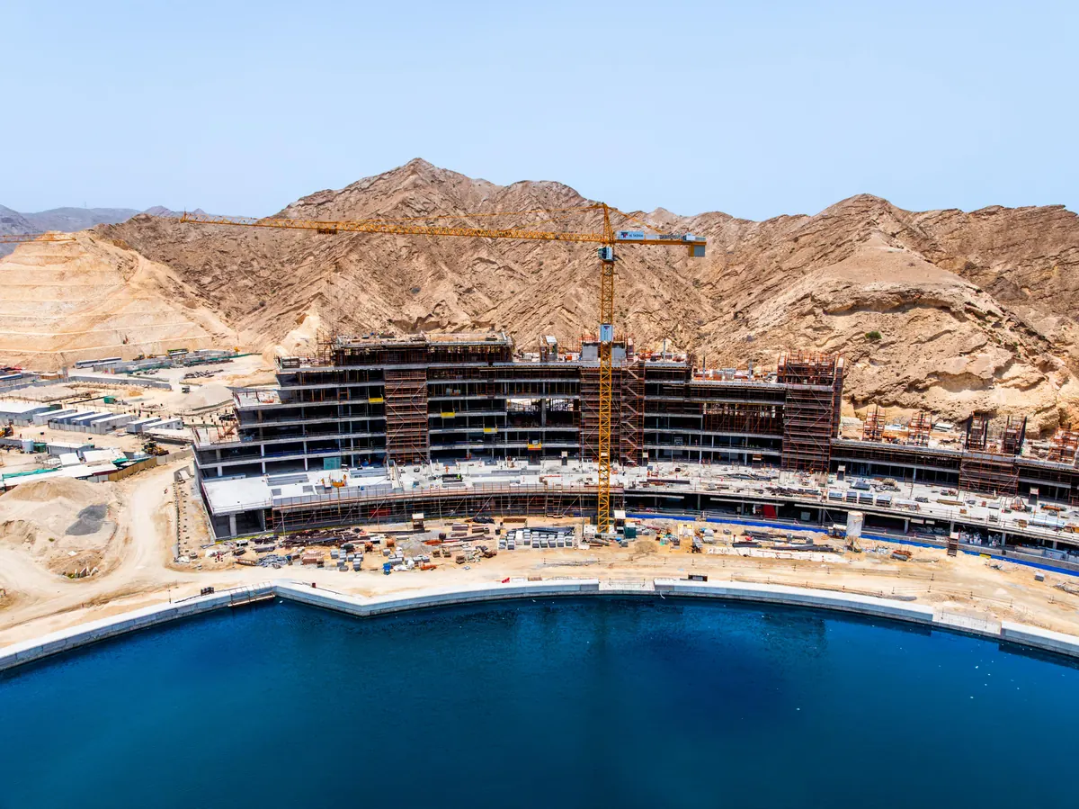 The structural frame of The Arc seen from the water, set against the Hajar mountains
