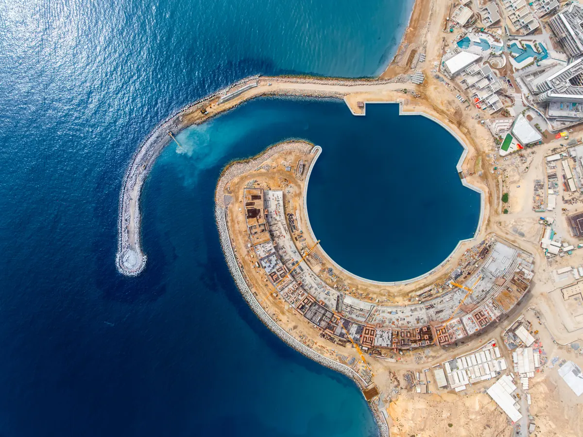 Aerial view directly above The Arc under construction in July 2026, the curved structure and the circular marina basin cut into the coastline